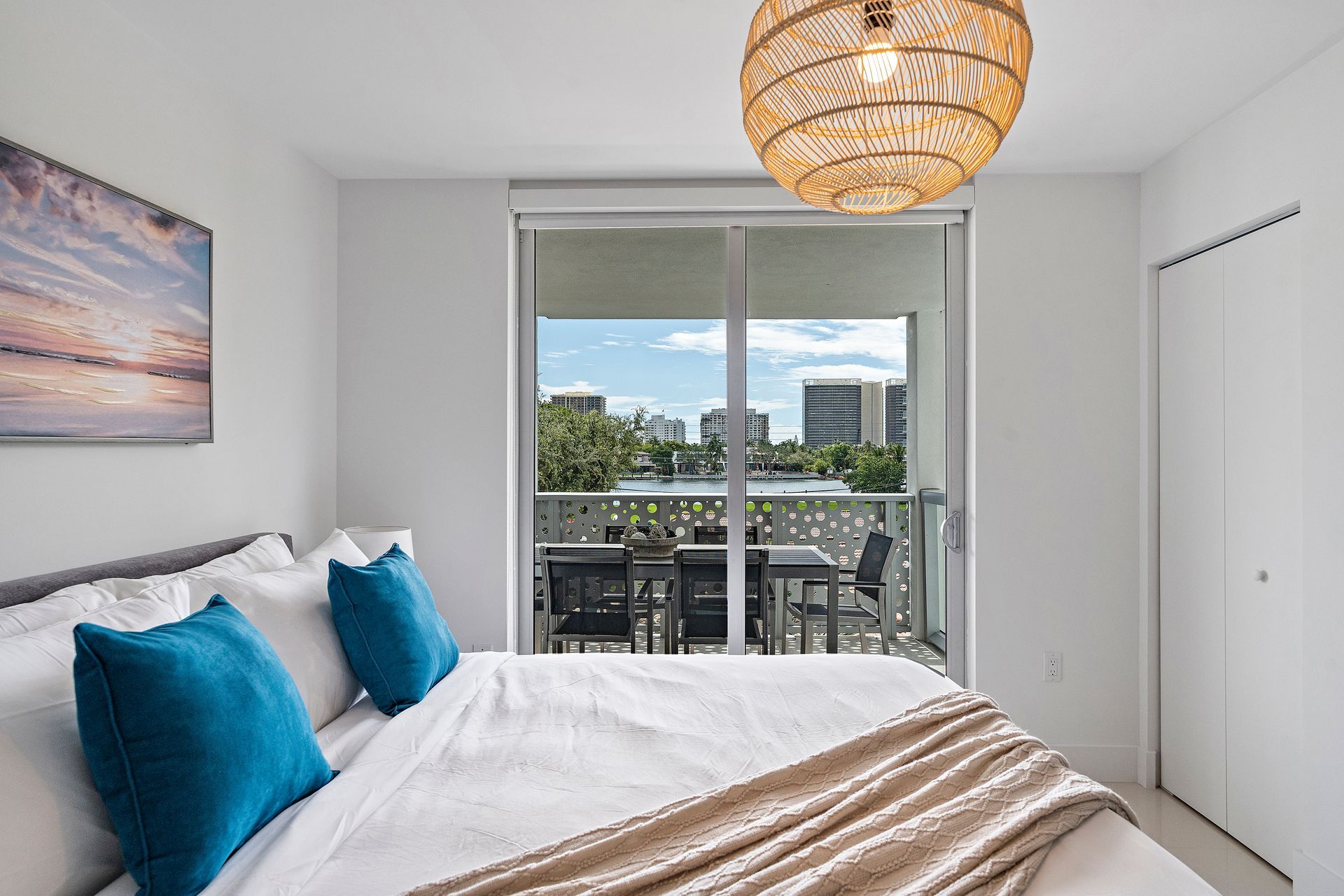 Bedroom with bed, balcony, and city view. Teal pillows, gold blanket, woven light fixture.