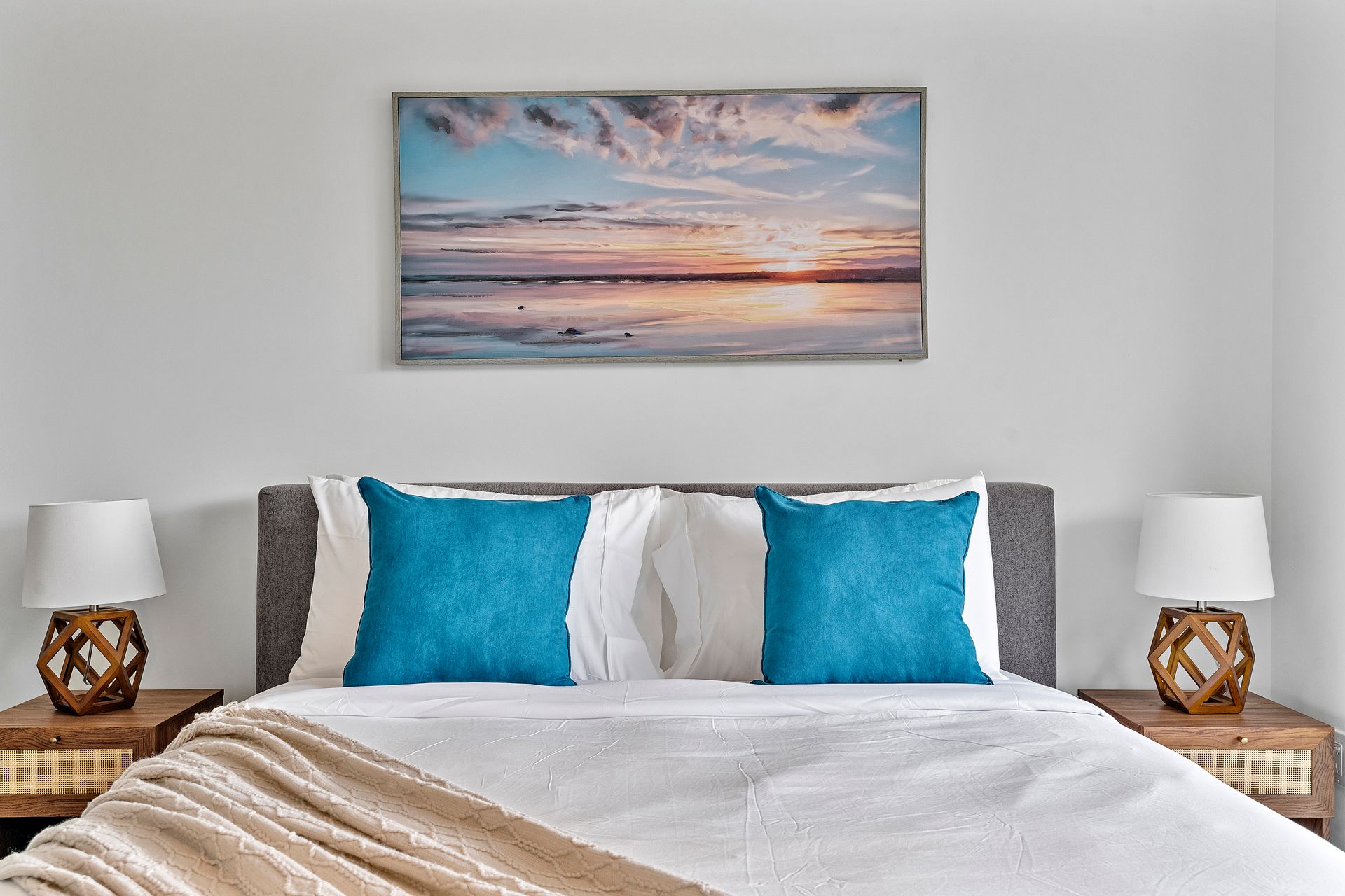 Bedroom with a bed, teal pillows, and a beach sunset painting above. Two lamps on nightstands.