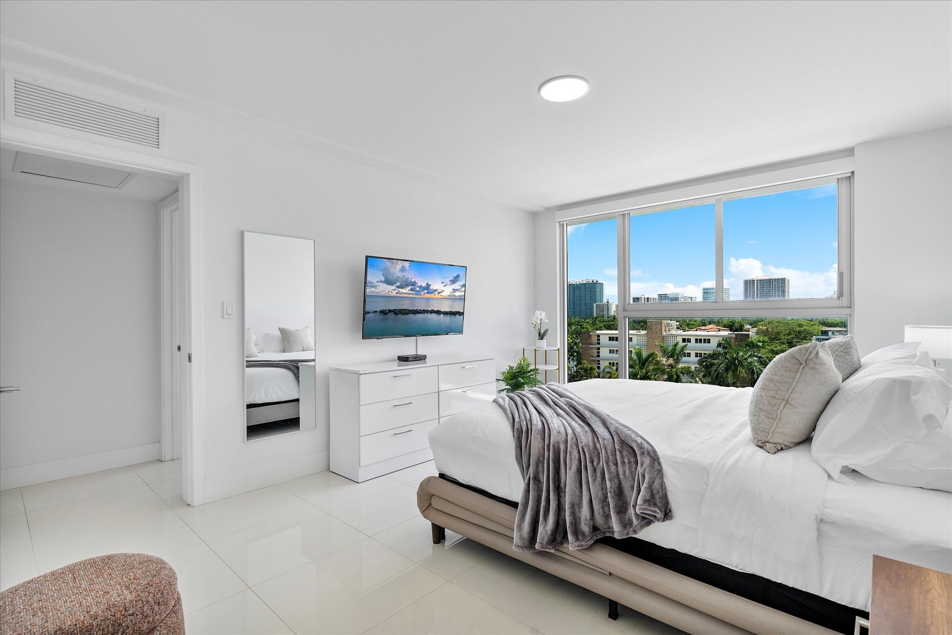 Bright white bedroom with large window overlooking a city skyline. Bed, dresser, TV, closet.