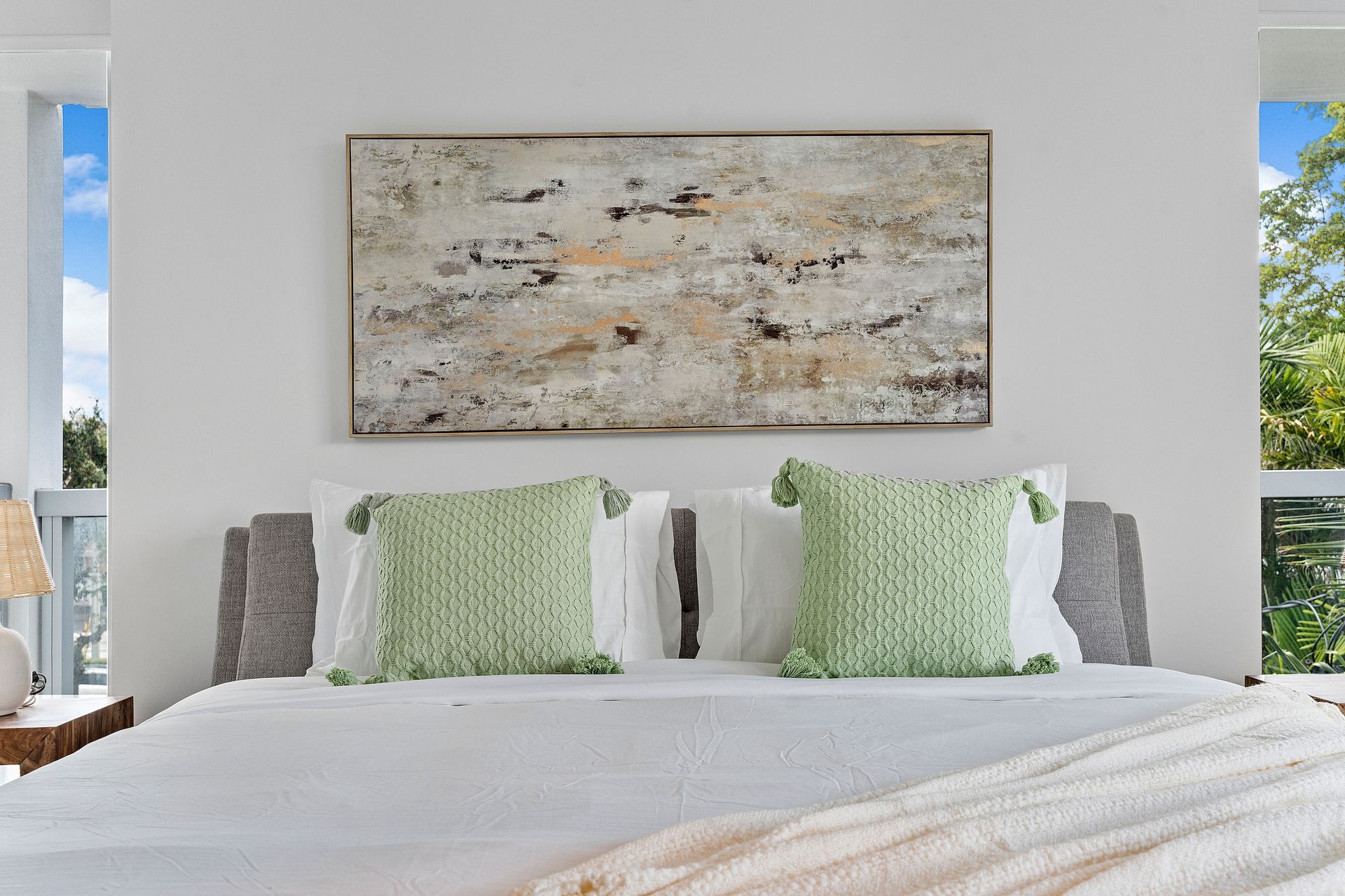 Bedroom with a bed, two green pillows, artwork, and windows with a view of greenery.