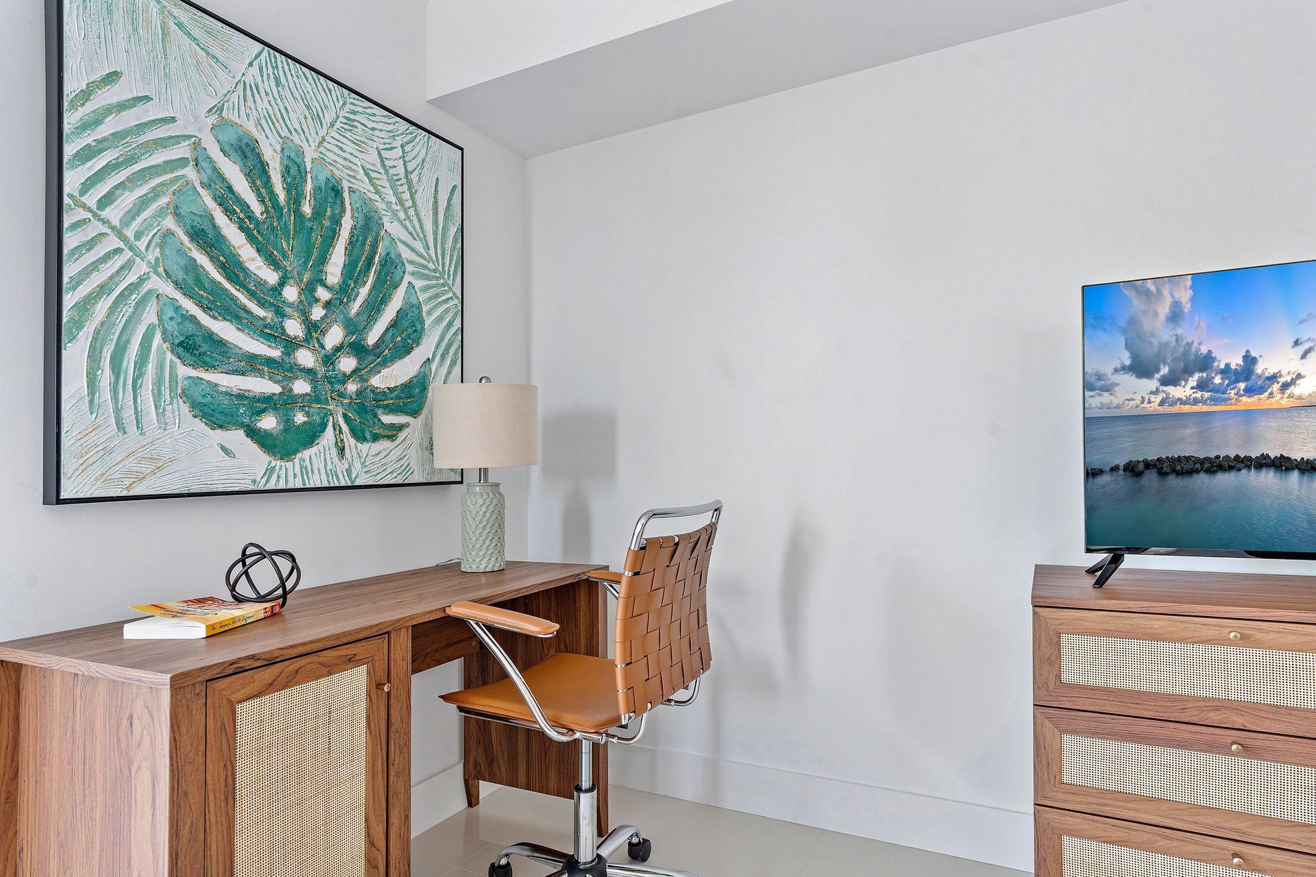 Desk in a bright room with a green leaf painting, lamp, chair, and TV showing ocean view.