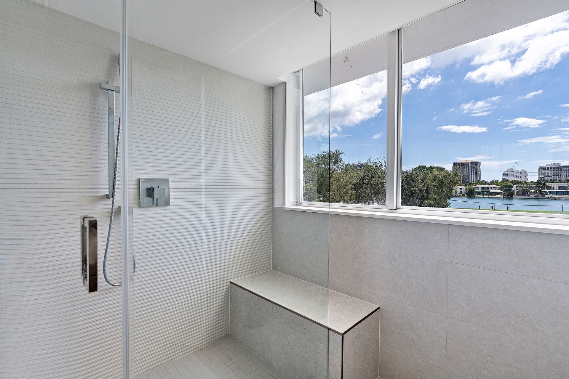 Modern white tiled shower with glass door, bench, and window overlooking a cityscape with blue sky.