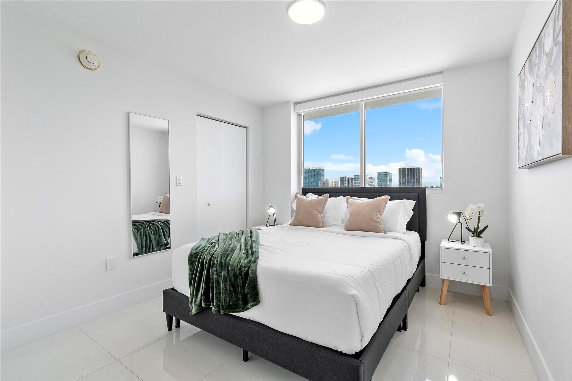 Bright bedroom with bed, large window, mirror, white walls and floor, city view.