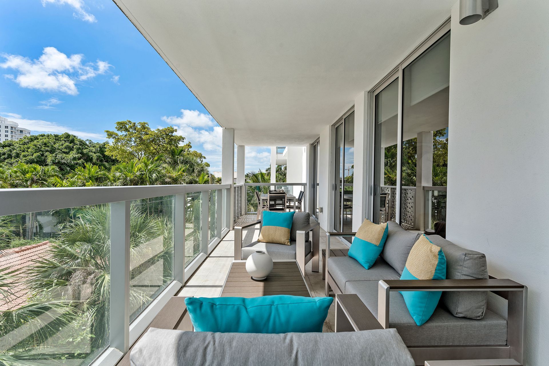 Balcony with gray sofa, turquoise pillows, and wooden tables, overlooking trees under a blue sky.