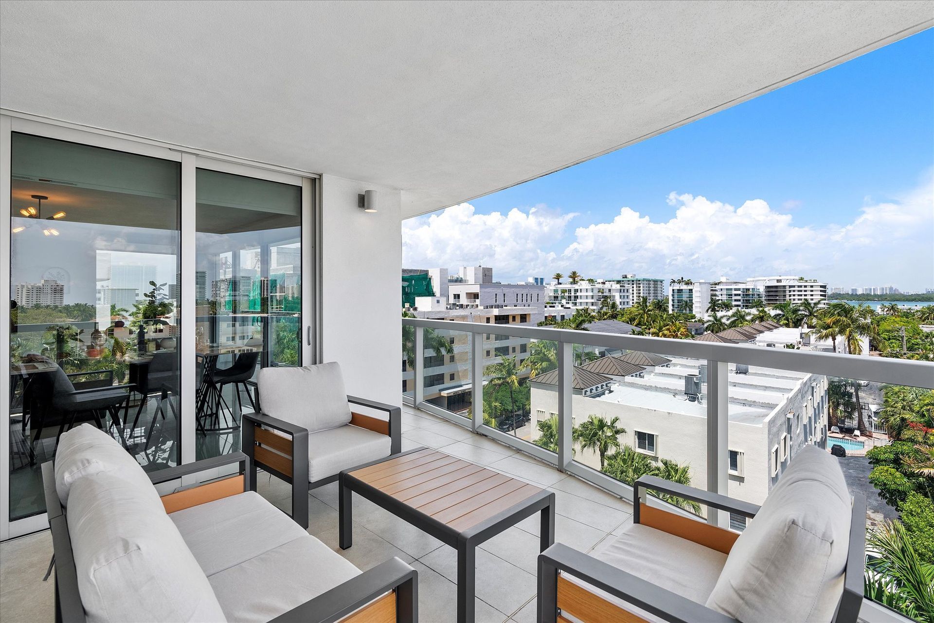 Balcony with modern furniture, cityscape view, and sliding glass doors. Blue sky visible.