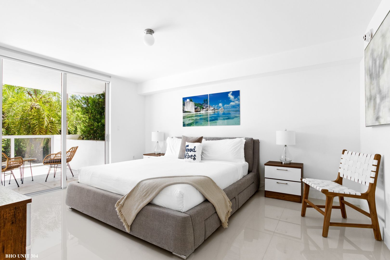Bright bedroom with a bed, balcony, and artwork; white walls, grey bed frame, beach scene art.