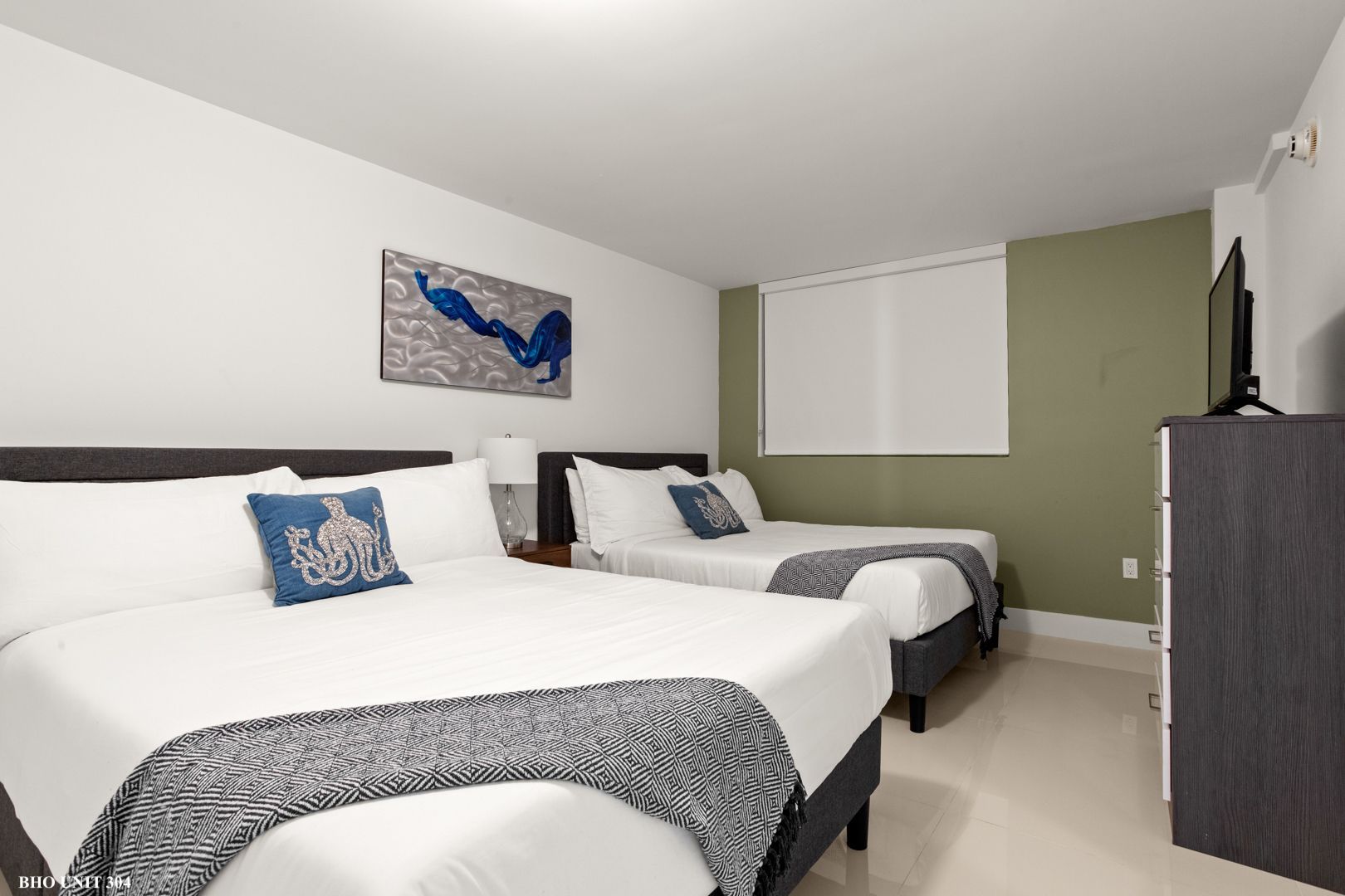 A hotel room with two beds, one king and one full. White bedding with blue and grey accents.