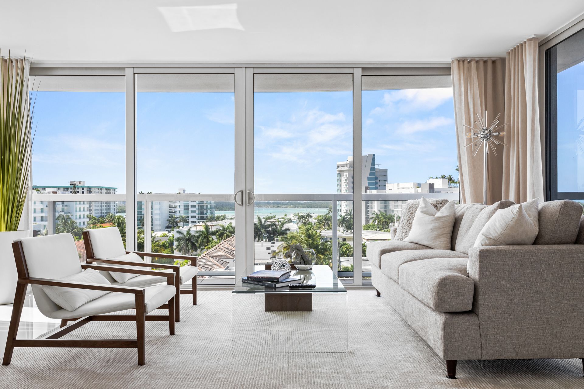 Living room with ocean view: grey sofa, white chairs, glass coffee table, and large sliding glass doors.