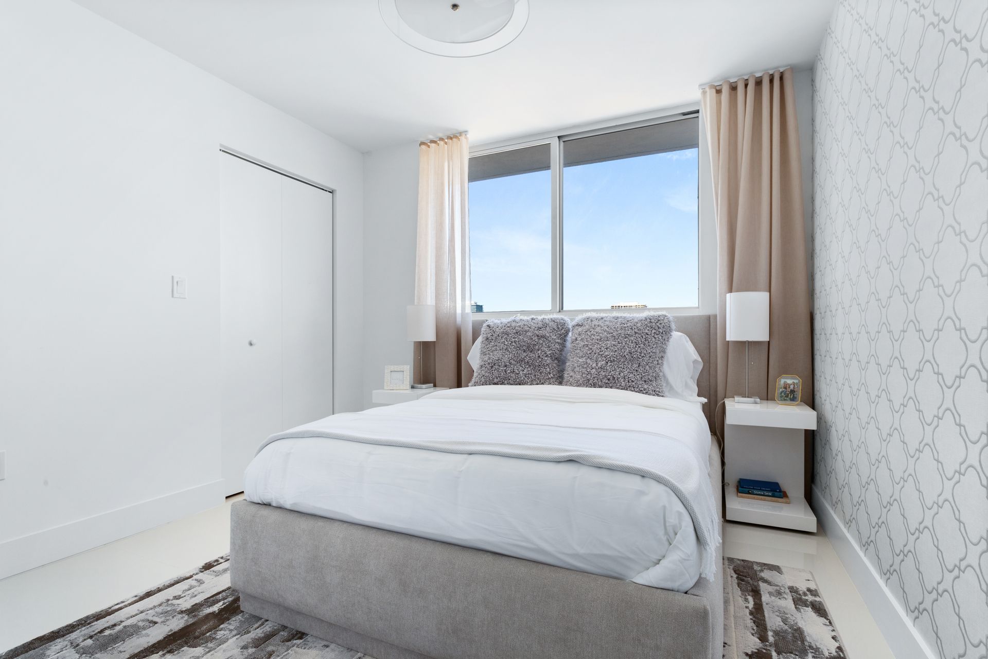 Bedroom with a bed, window, white walls, rug, and beige curtains.