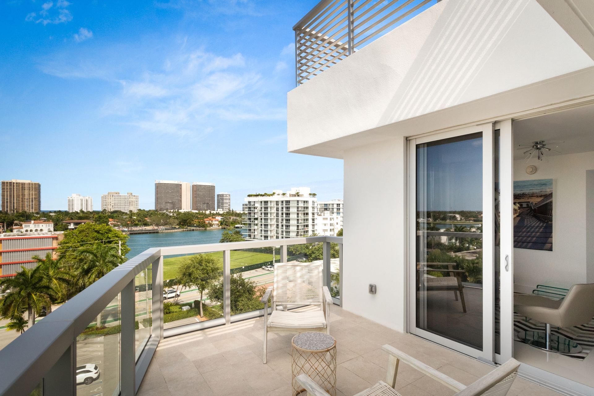 Balcony with clear railing, white chairs, and city view on a sunny day.
