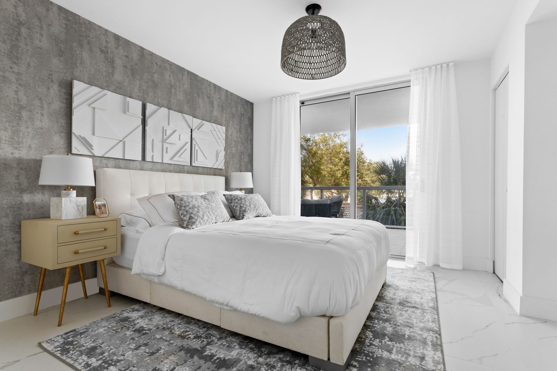 Bedroom with a white bed, gray textured wall, gold bedside tables, and a balcony view.