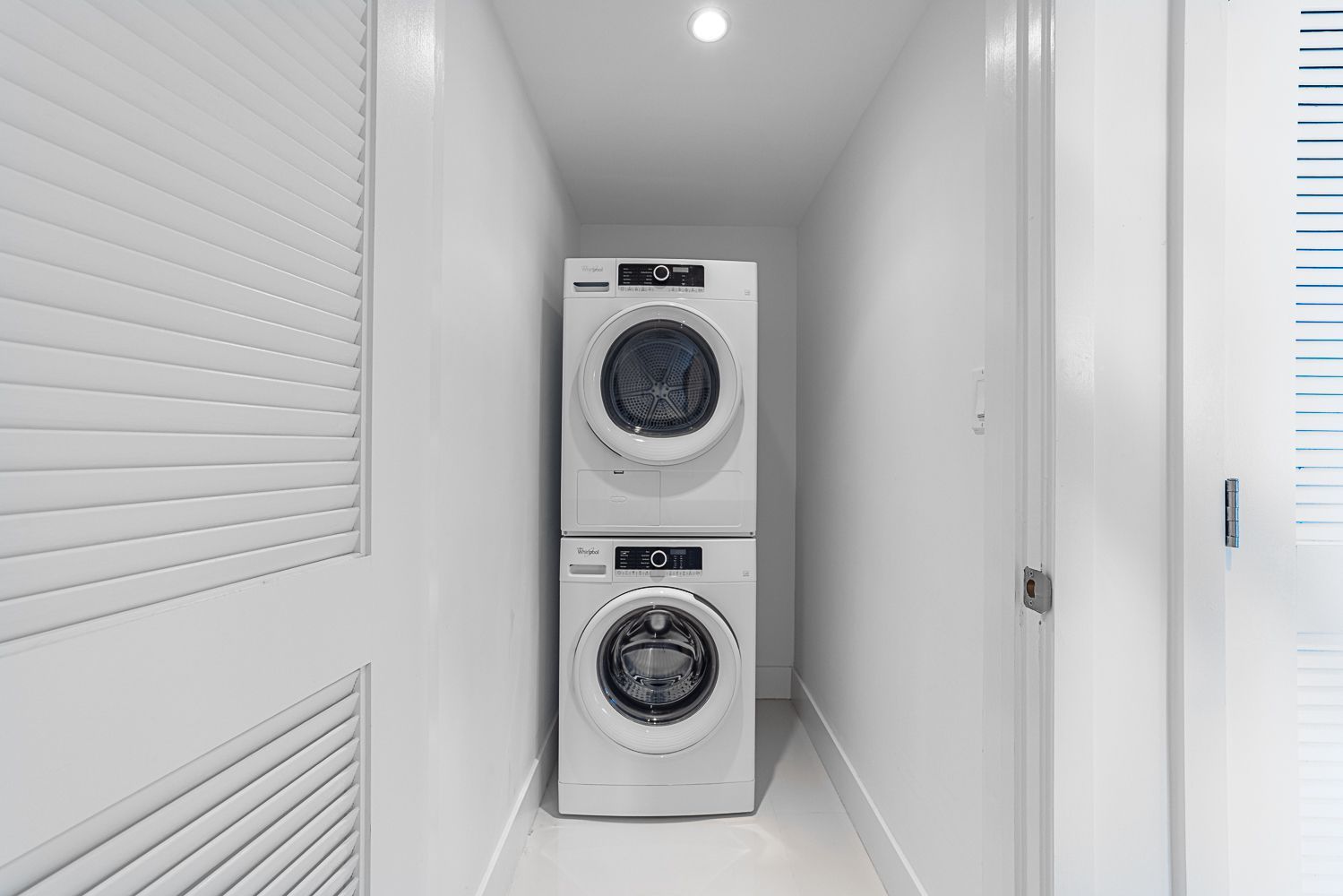 Stacked white washing machine and dryer in a narrow, white hallway with a door and slatted window.