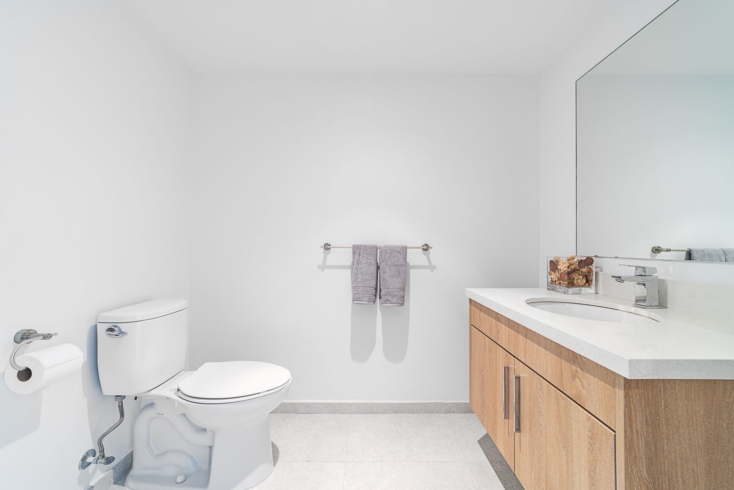 White bathroom with toilet, vanity, mirror, and towel rack; towels are gray.