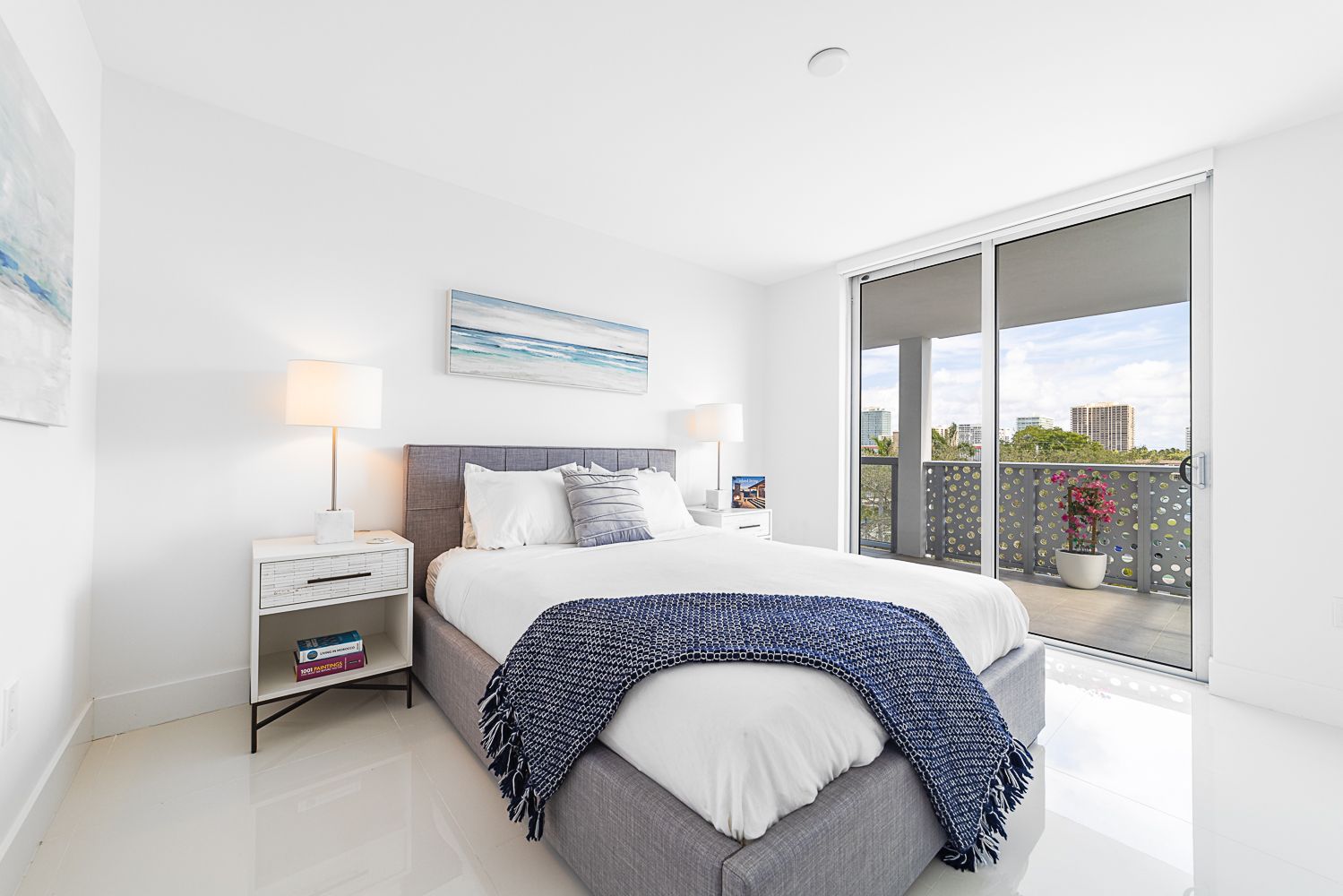 Bright bedroom with bed, nightstands, balcony, and ocean-themed artwork.