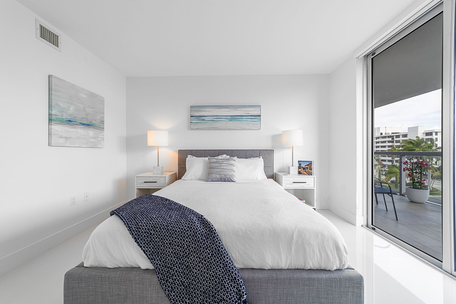 Bedroom with white walls, grey bed, balcony view. Blue and white art.