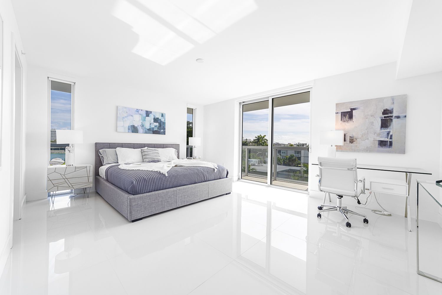 Bright, white bedroom with bed, desk, and balcony. Artwork and large windows, minimalist design.