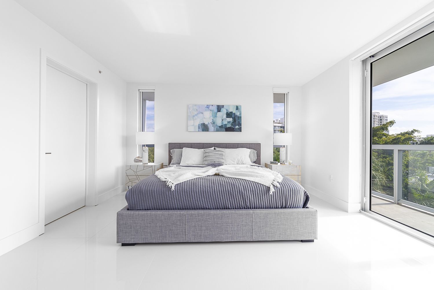 White bedroom with gray bed, artwork, and large window overlooking a balcony.