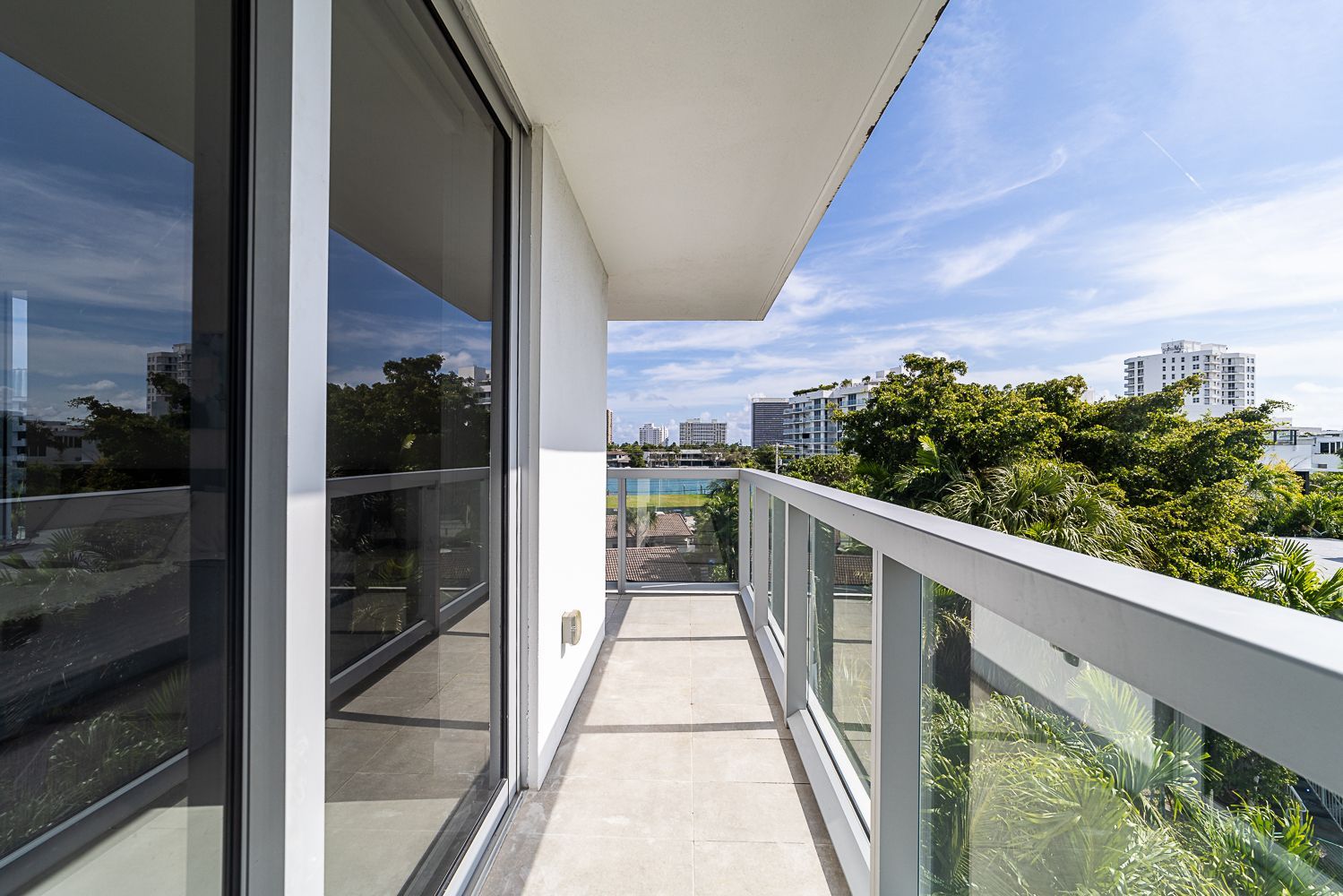 Balcony with glass railing, overlooking buildings and trees under a blue sky.