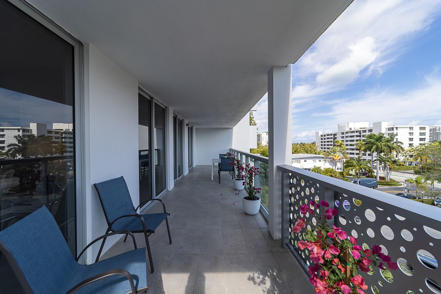 Balcony with blue chairs, white walls, and a metal railing with flowers; overlooking a sunny city scene.