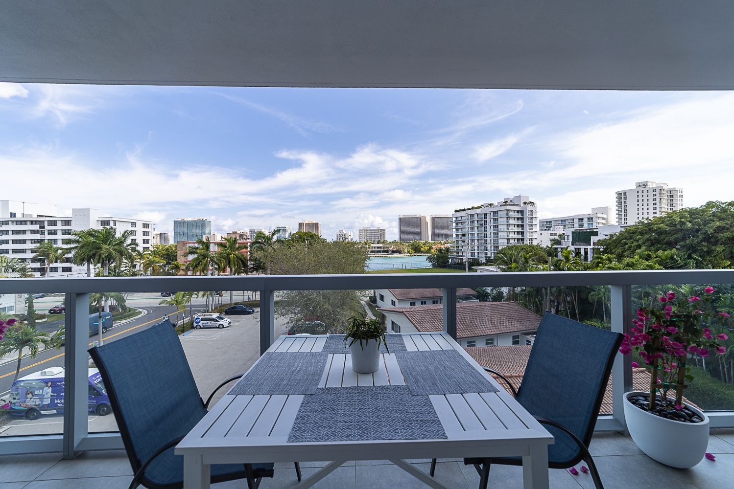 Balcony with table and chairs, overlooking a city with buildings and a blue sky.