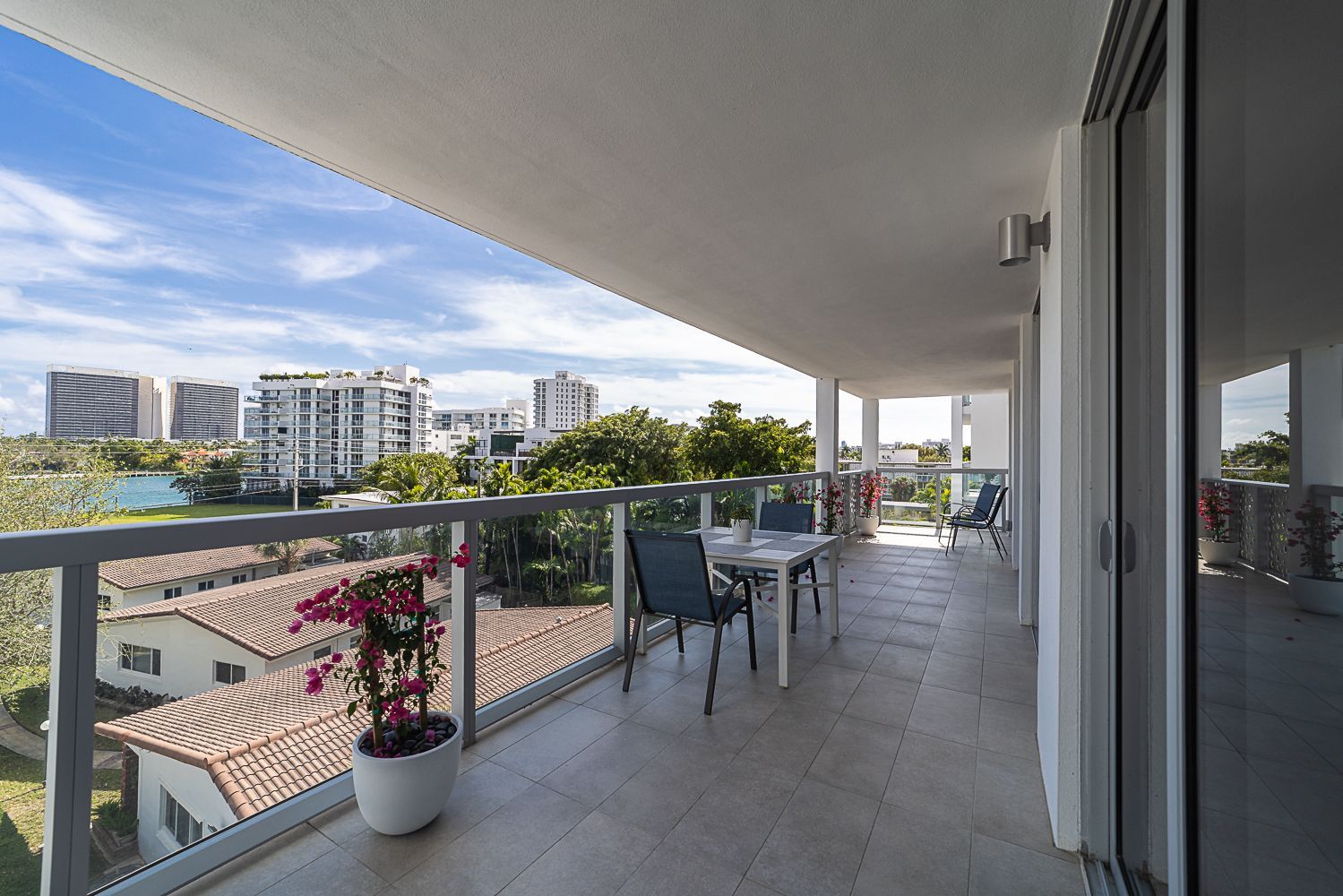 Balcony with table, chairs, and city views. Clear glass railing. Flowers in a pot. Sunny day.