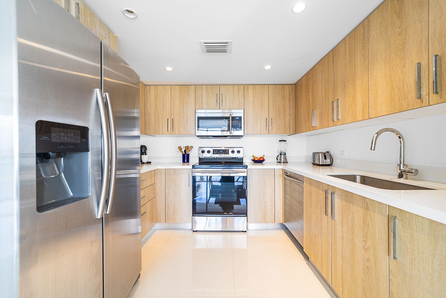 Modern kitchen with stainless steel appliances, light wood cabinets, white countertops, and tile flooring.