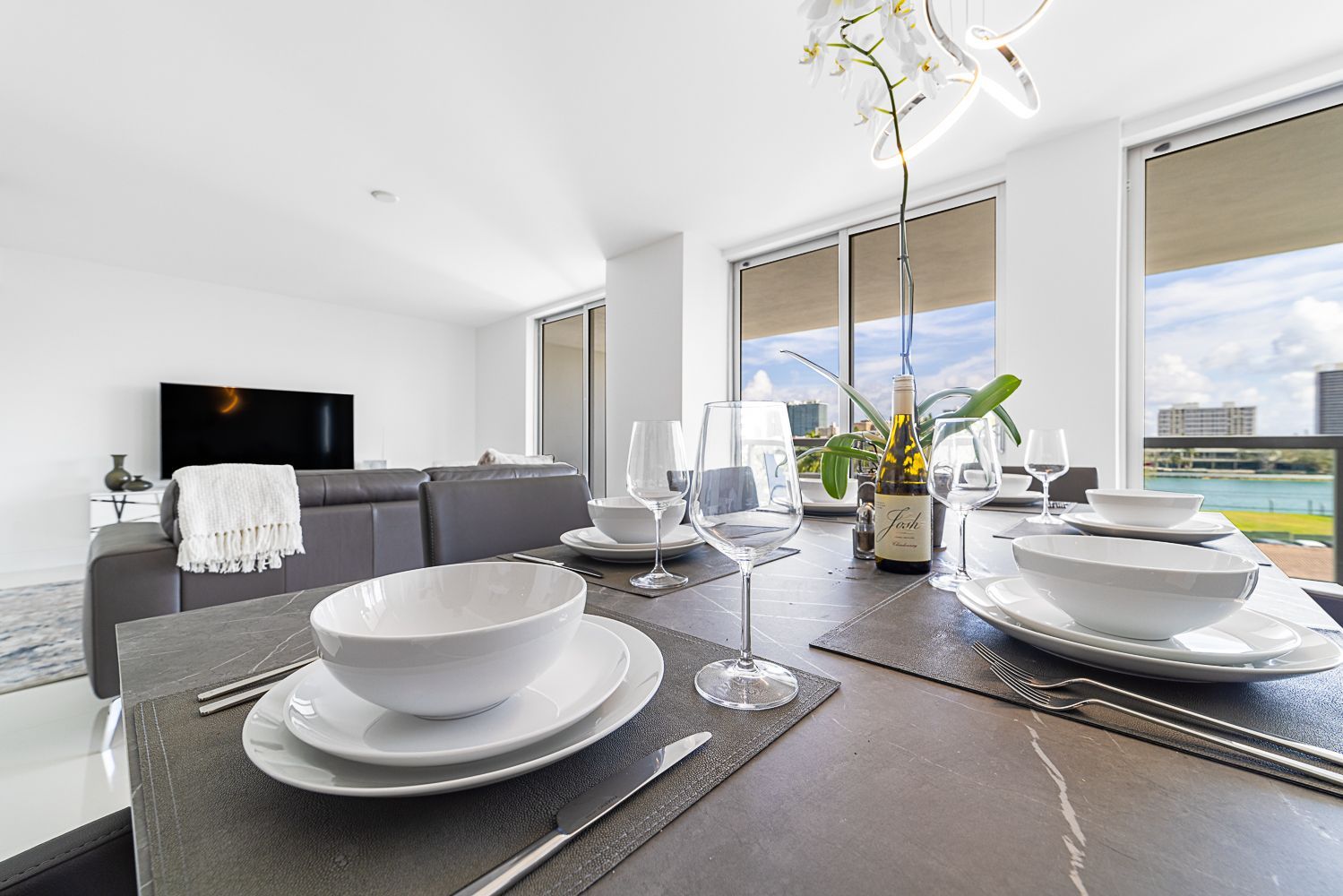 Dining table set with white tableware, city view through windows. Living room with gray sofa and TV visible.