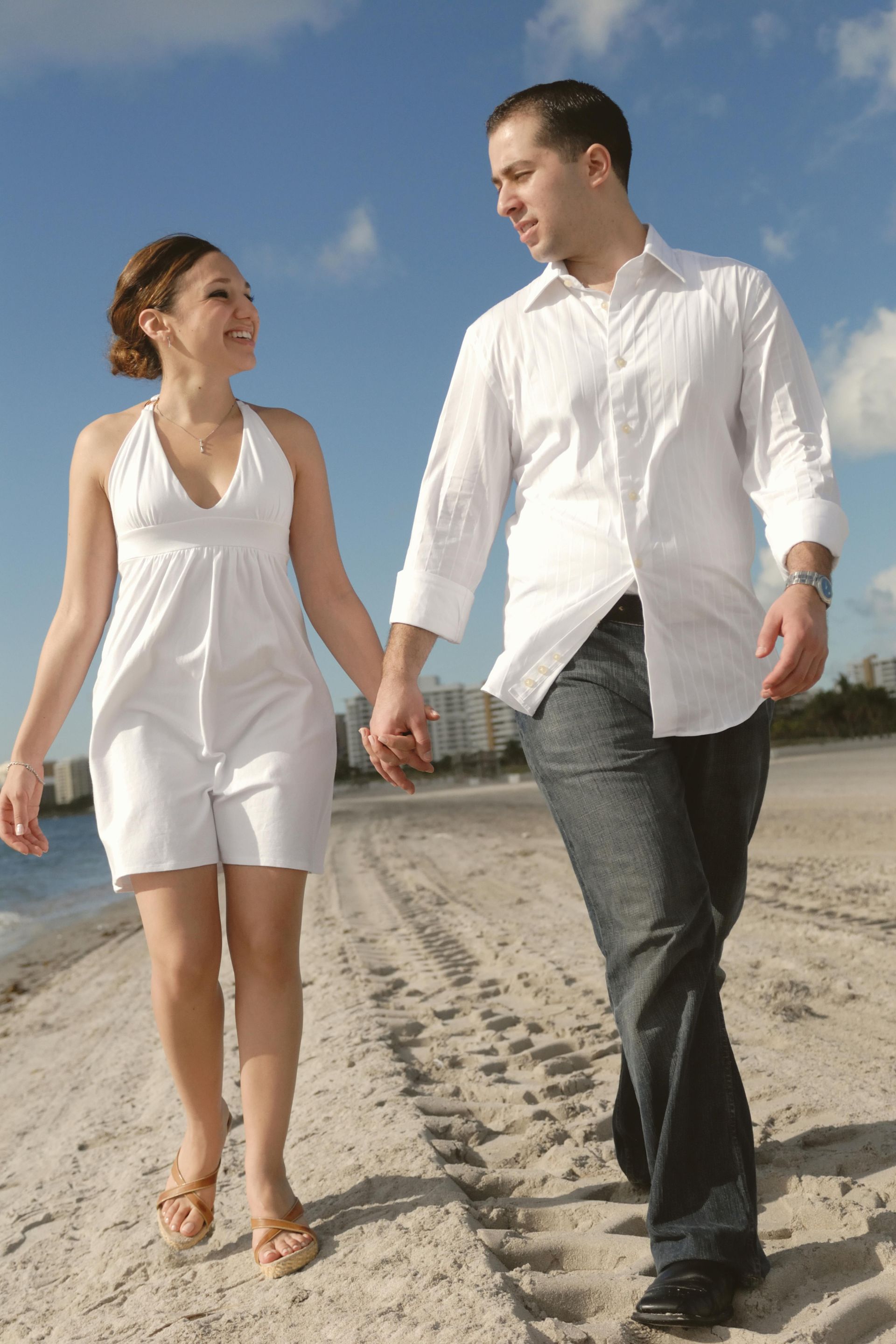 Man and woman laughing together outdoors, leaning against building. Man in black shirt, woman in white, with green cap.