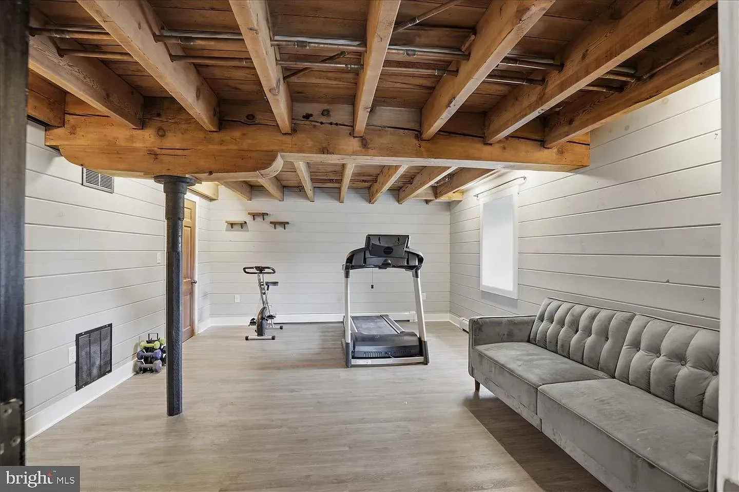 Basement home gym with exposed wooden beams, treadmill, exercise bike, and gray couch against white walls