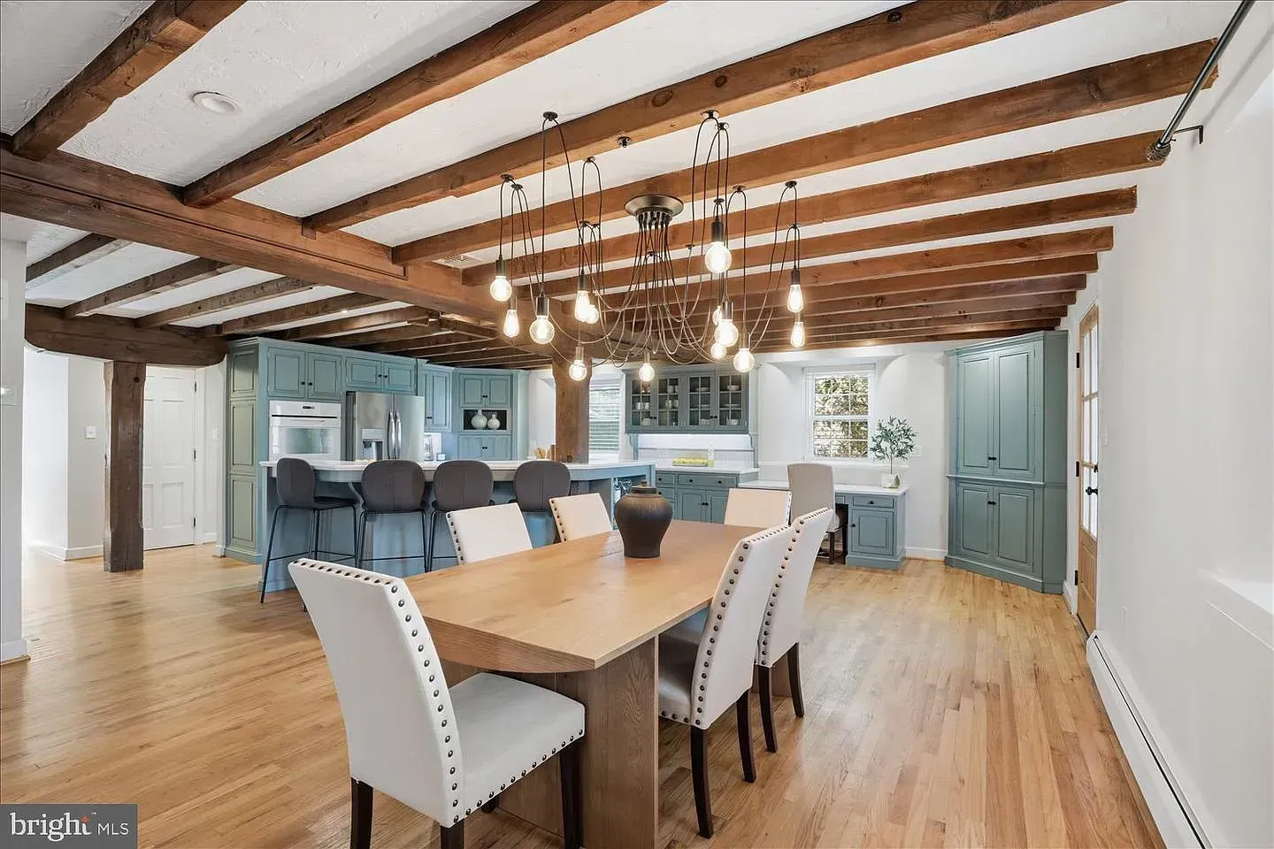 Bright dining room with exposed beams, chandelier, large table, white chairs, and blue kitchen cabinets