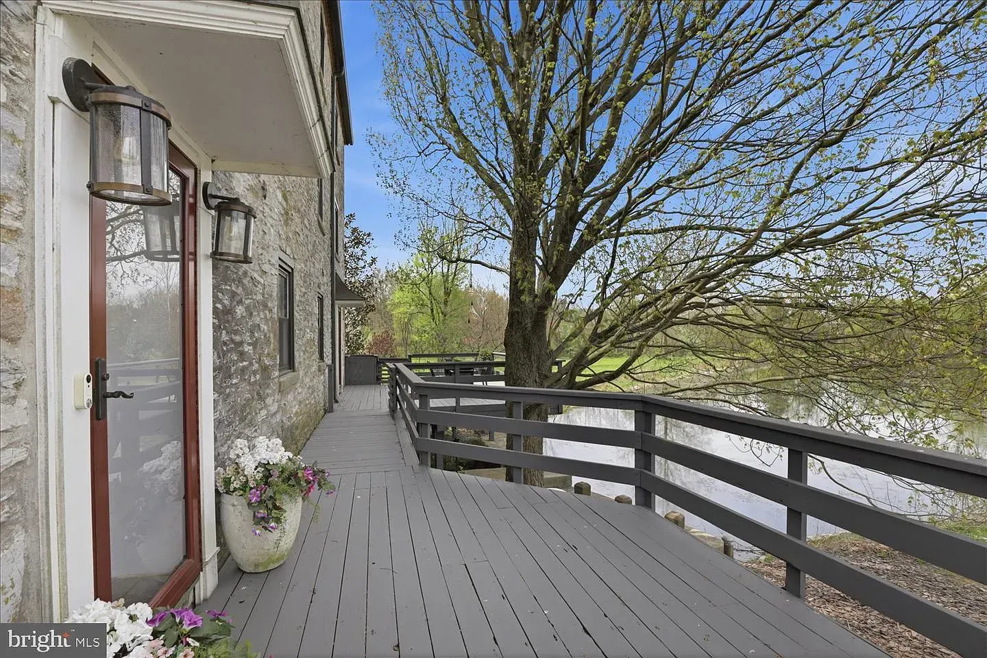 Wooden deck beside a stone house, overlooking a river with a leafless tree and railing.
