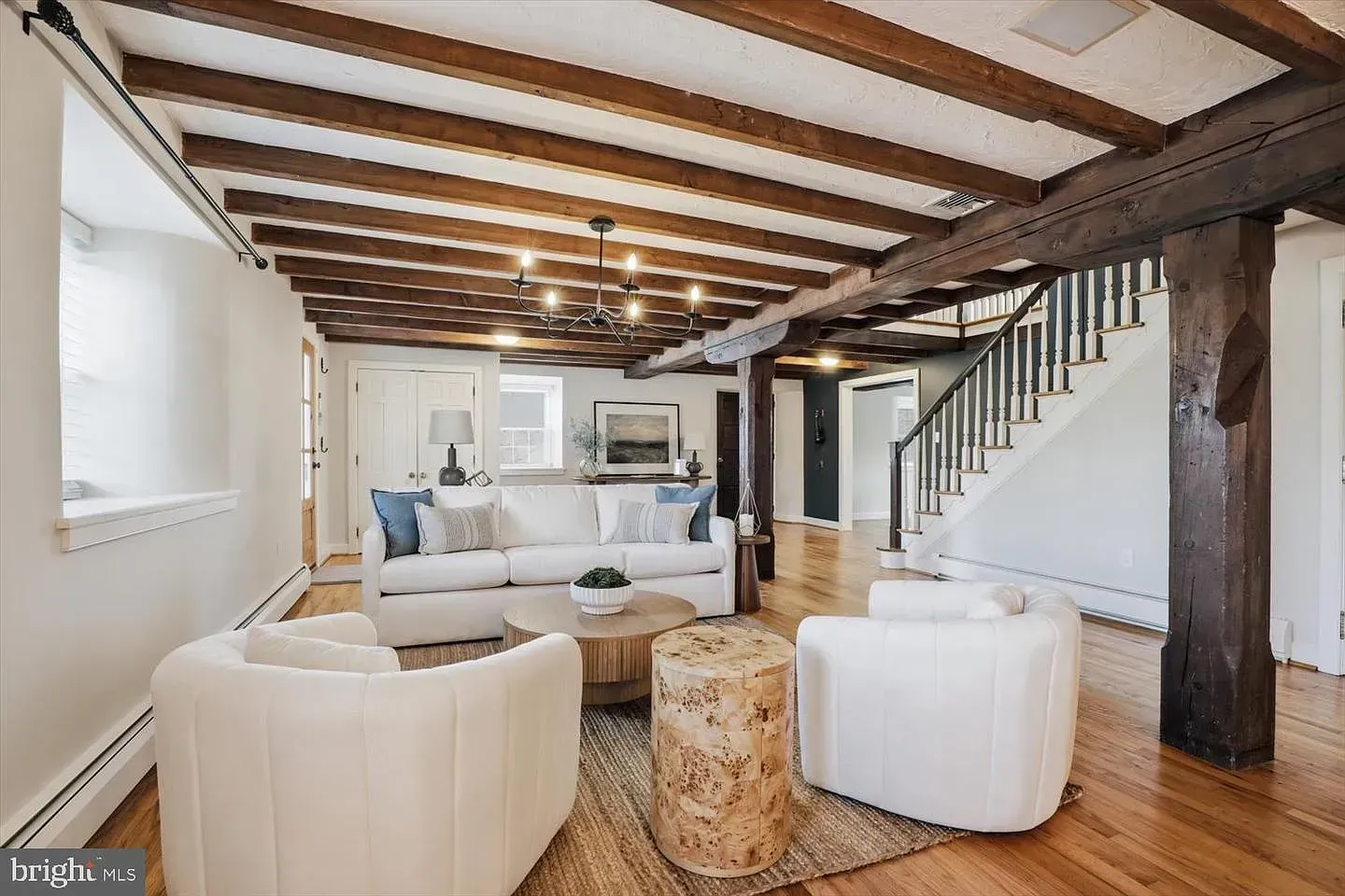 Bright living room with exposed wooden beams, white furniture, staircase, and patterned rug