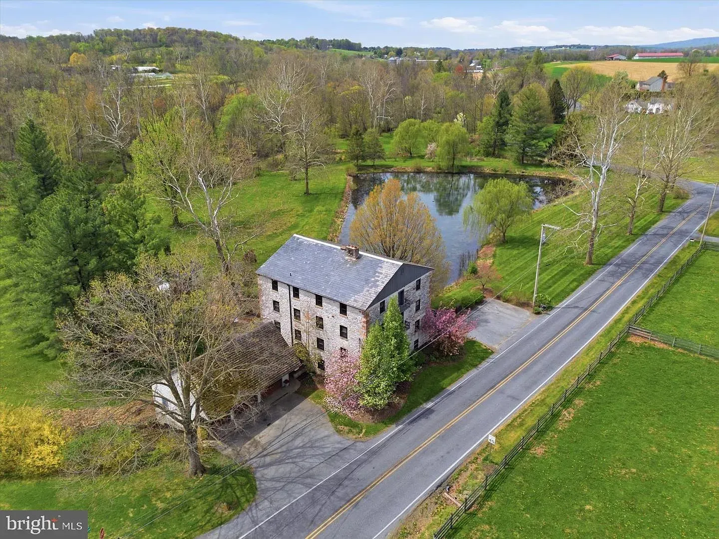 Aerial view of a rural stone house beside a road, with a pond and green fields in the background