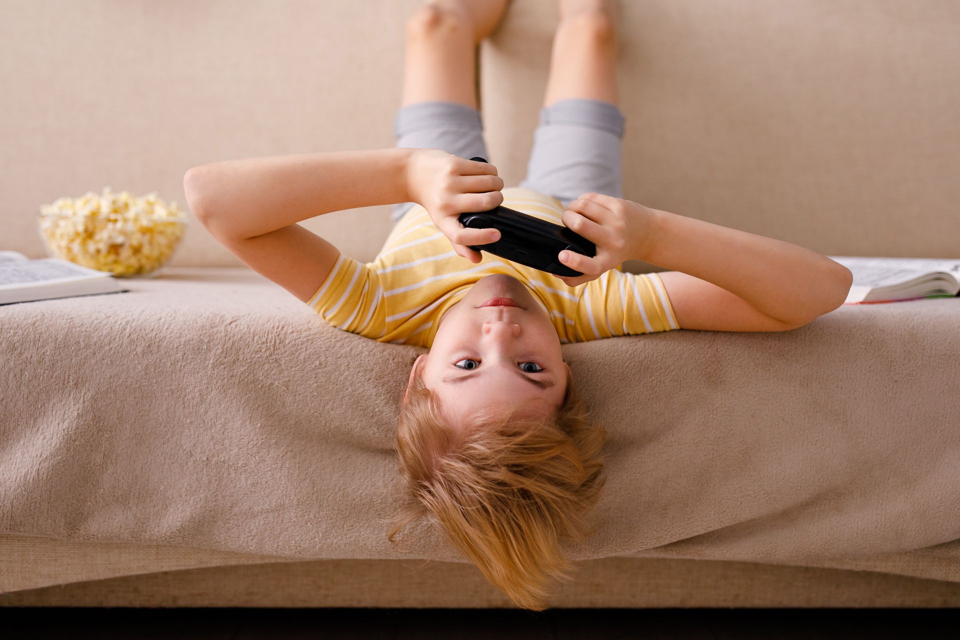 Boy on lounge upside down, holding gaming controller with popcorn next to him.