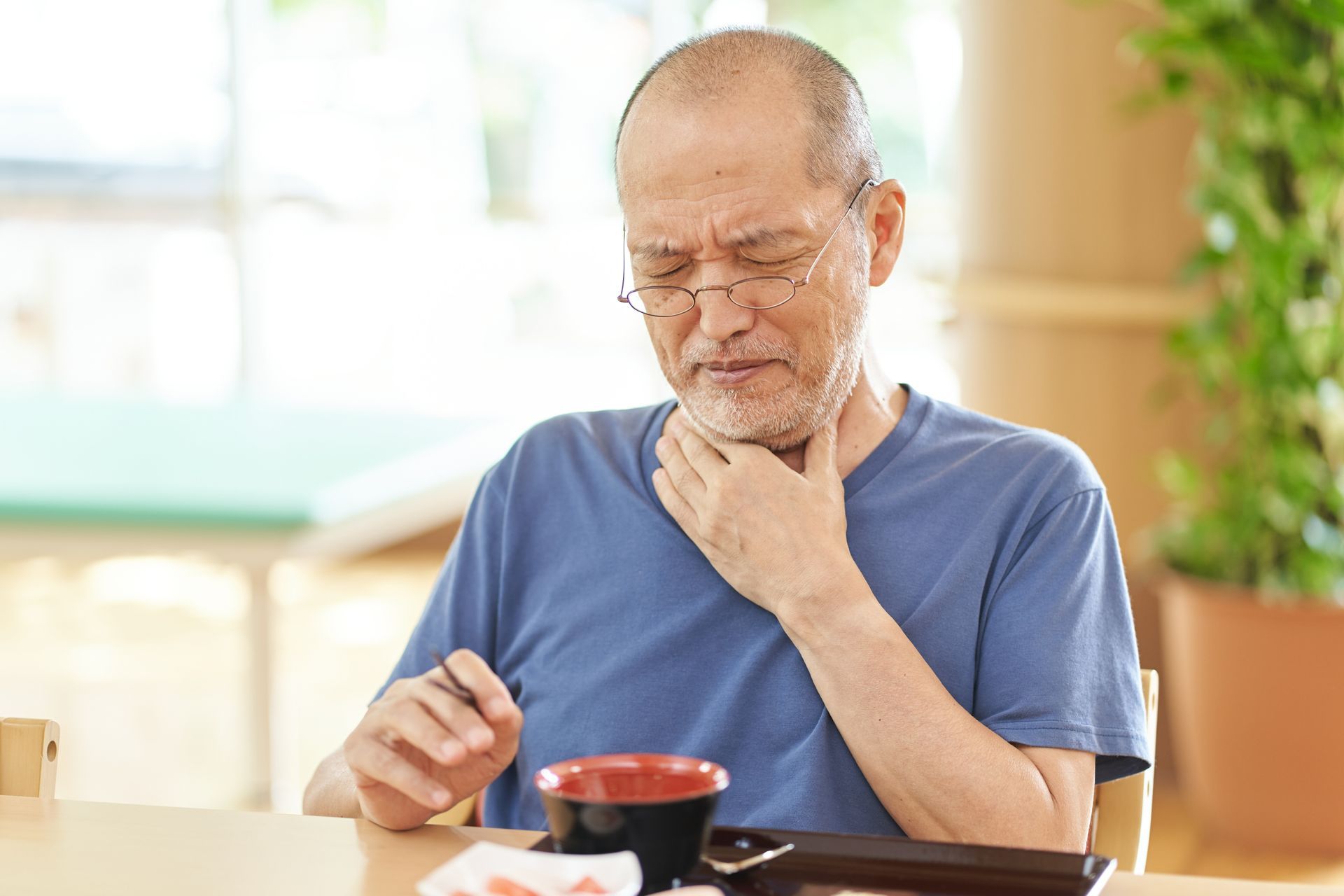 An old man sitting at a table holding his throat, looking to be struggling to swallow