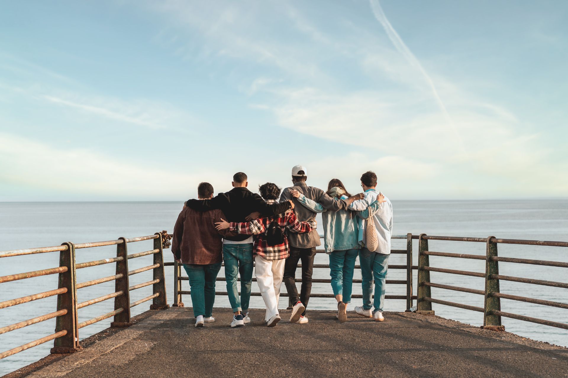 Group of diverse young people standing on jetty looking out at water arms around each other.