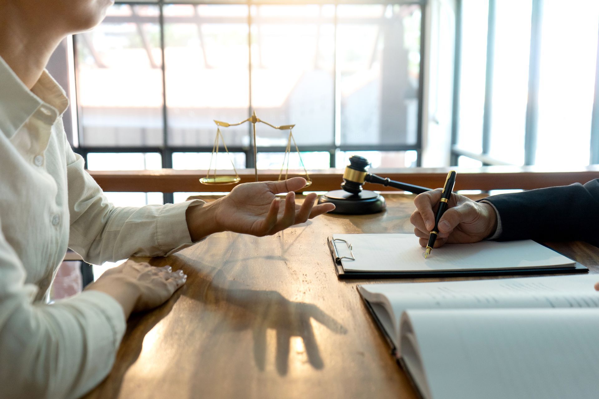 Person gesturing while consulting with a lawyer at a desk in an office; scales of justice and gavel in background.