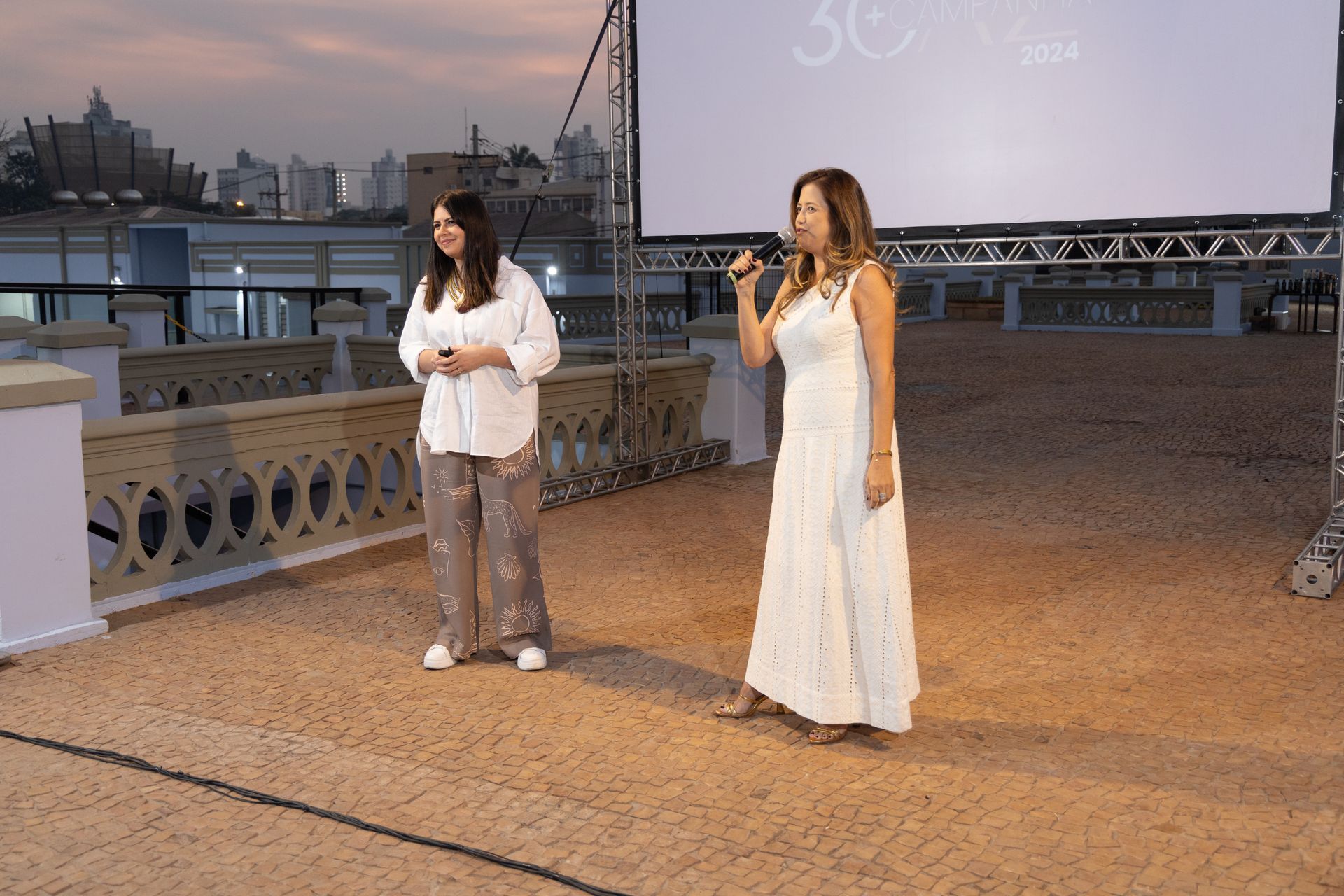 Duas mulheres em vestidos brancos estão em pé na frente de uma tela grande.