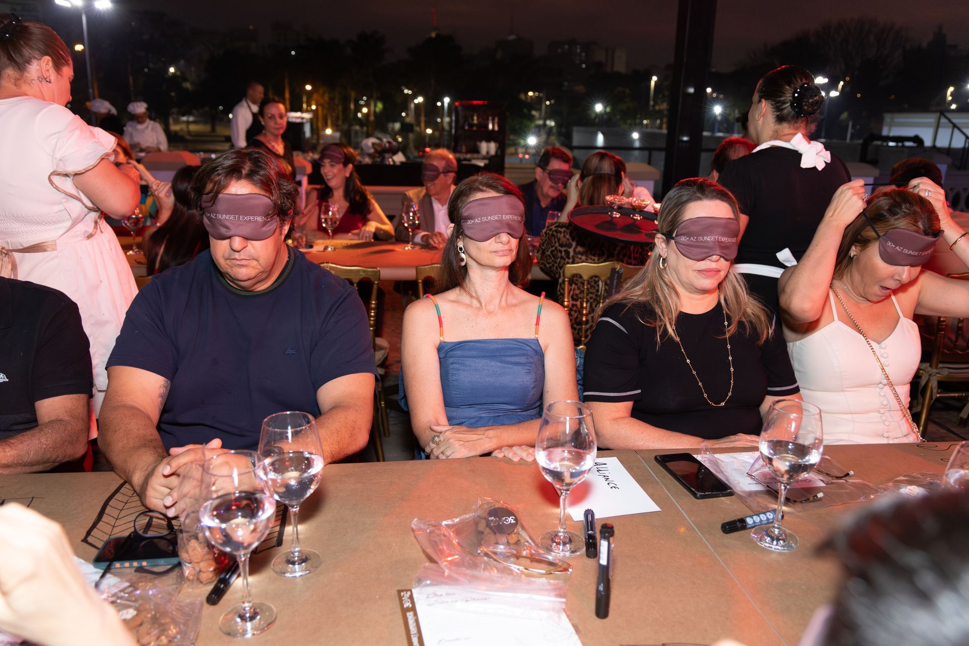 Um grupo de pessoas usando vendas nos olhos estão sentadas à mesa com taças de vinho.