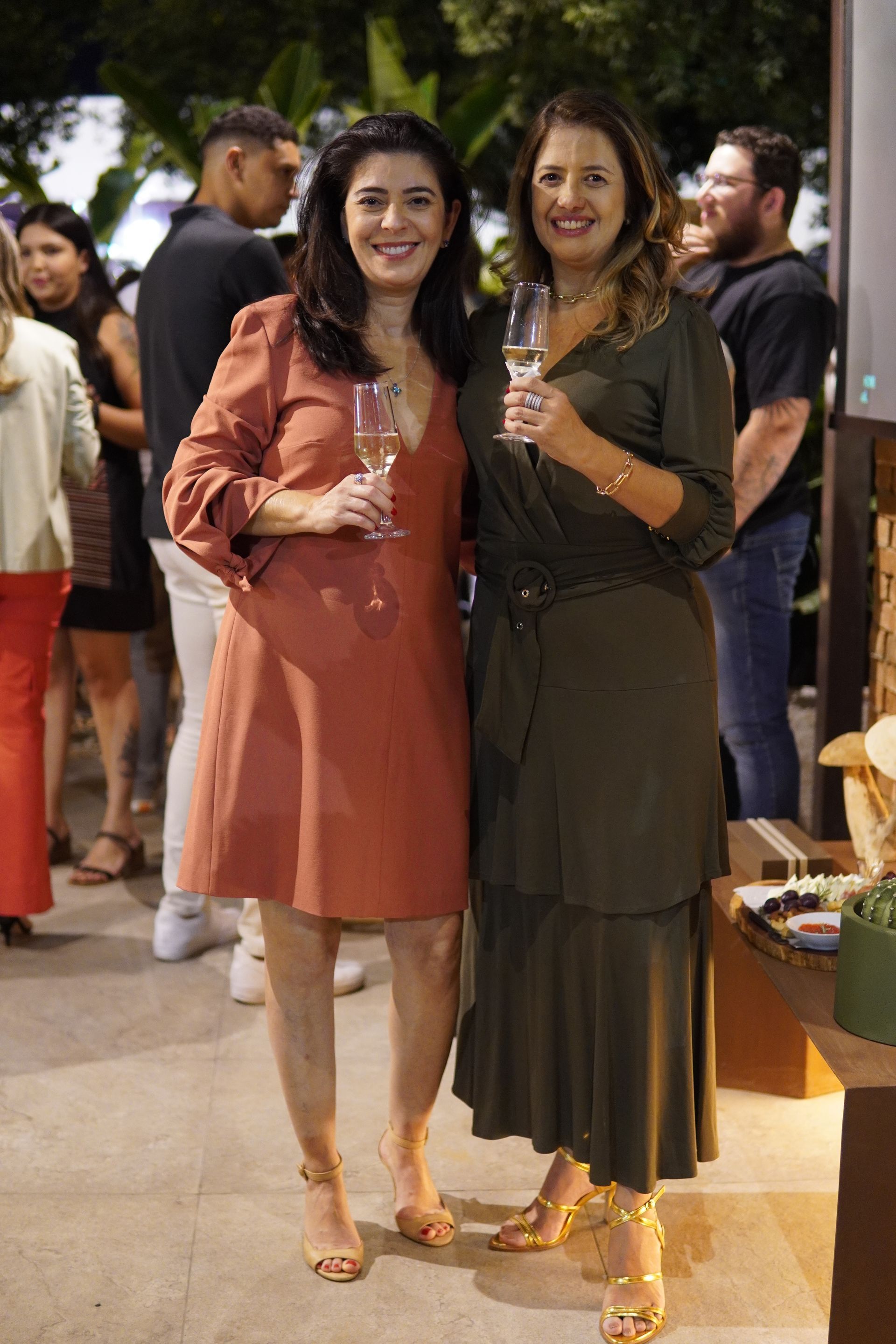 Duas mulheres estão de pé, uma ao lado da outra, segurando taças de vinho.