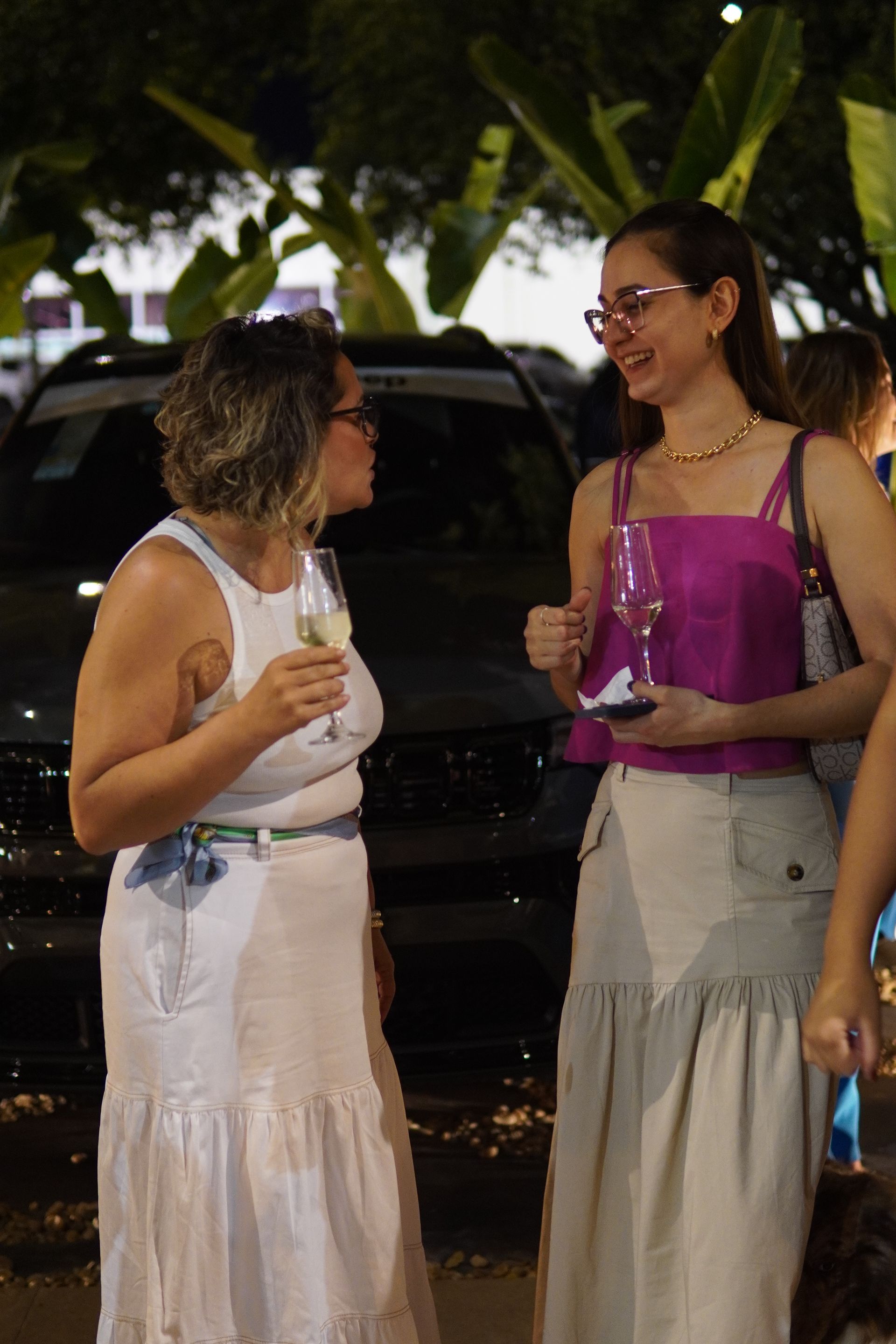 Duas mulheres estão de pé, uma ao lado da outra, segurando taças de vinho.