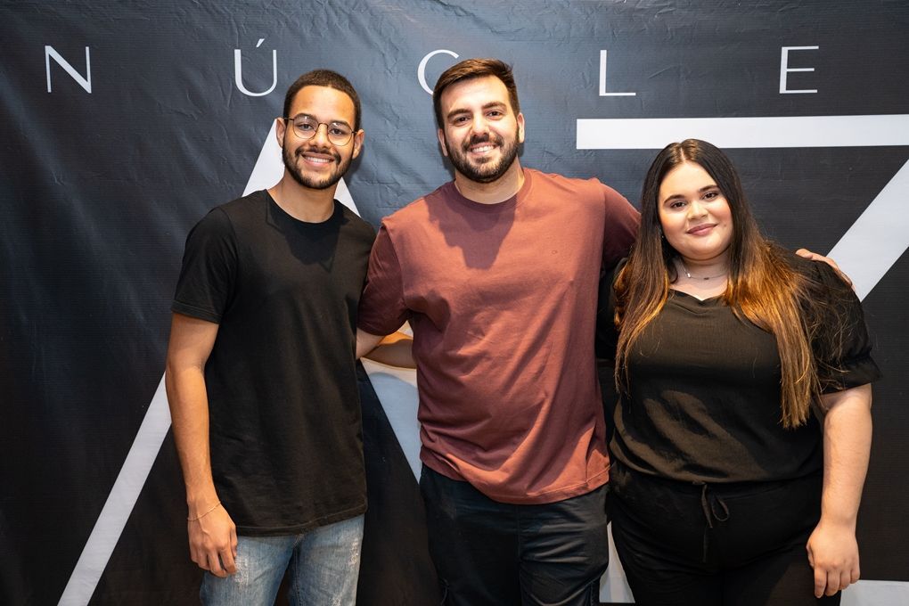 Três pessoas estão posando para uma foto em frente a um banner.