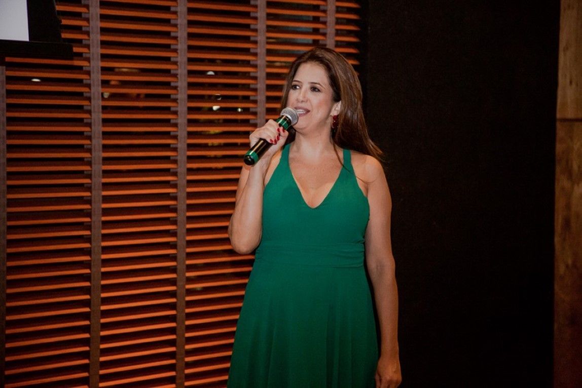 Uma mulher de vestido verde segura um microfone em frente a uma parede de madeira.