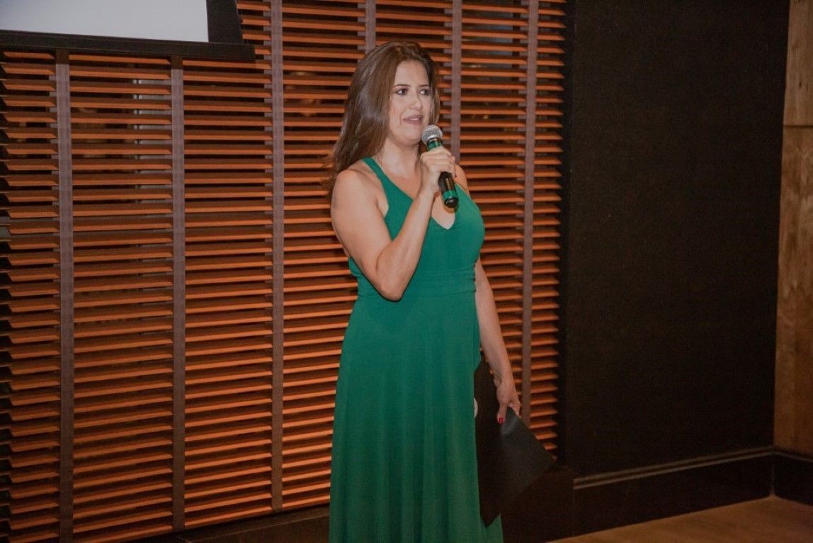 Uma mulher de vestido verde segura um microfone em frente a uma parede de madeira.