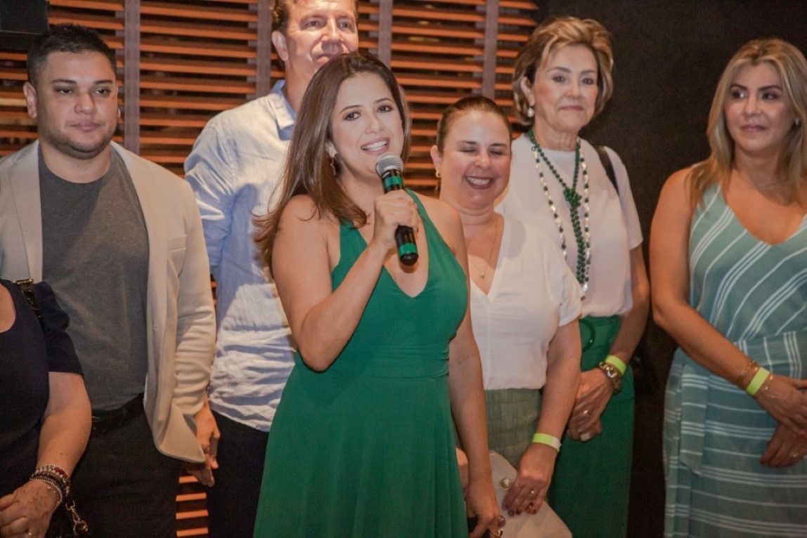 Uma mulher de vestido verde segura um microfone na frente de um grupo de pessoas.