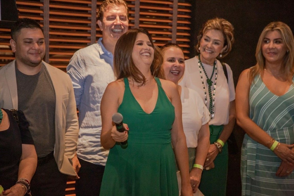 Uma mulher de vestido verde segura um microfone na frente de um grupo de pessoas.