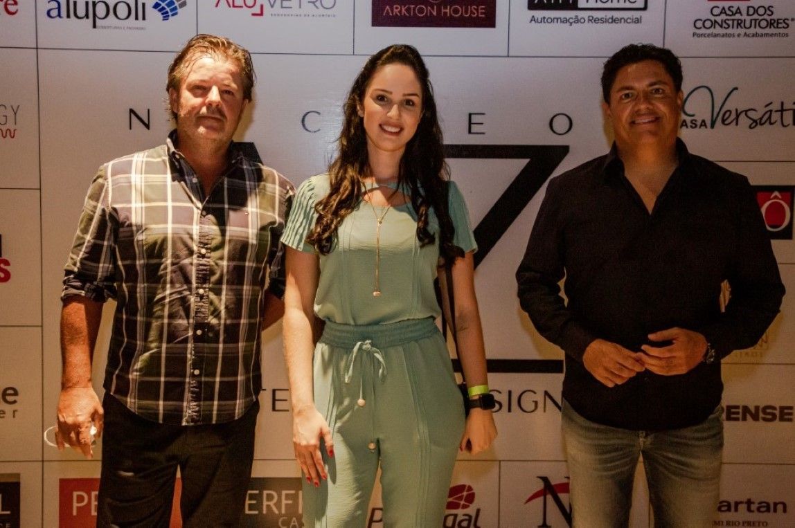 Três pessoas estão posando para uma foto em frente a uma parede com logotipos.