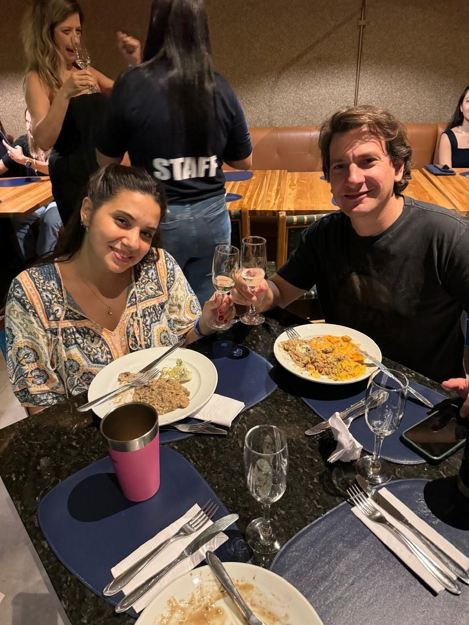 Um homem e uma mulher estão sentados à mesa com pratos de comida e taças de vinho.