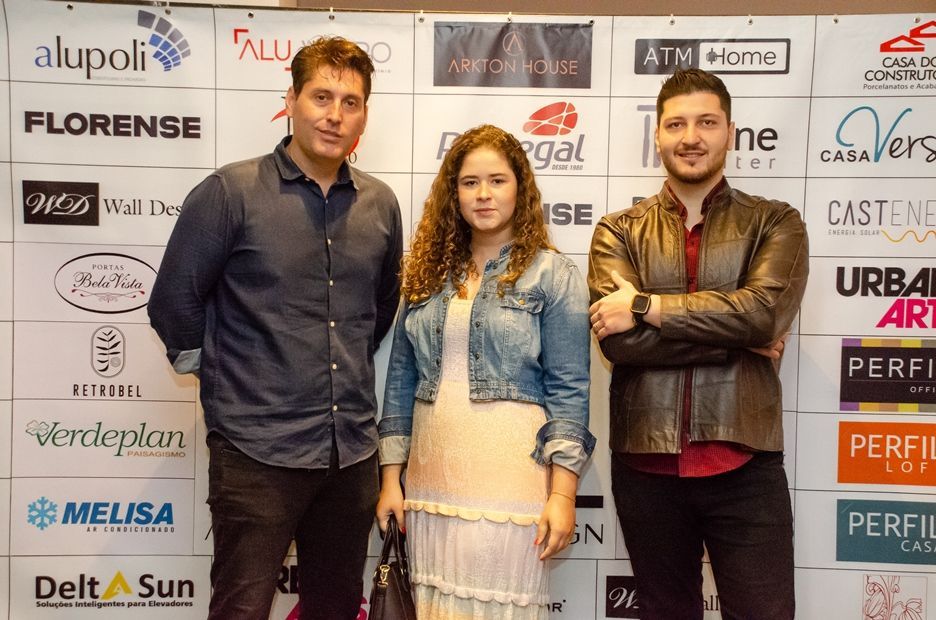 Três pessoas estão posando para uma foto em frente a uma parede com logotipos.