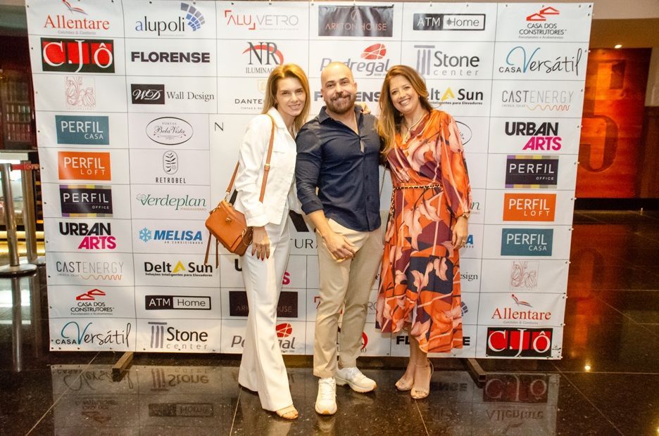 Três pessoas estão posando para uma foto em frente a uma parede com logotipos.